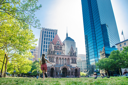 Boston Trinity Church with skyscrapers during summer day, with a teenager girl running after pigeons