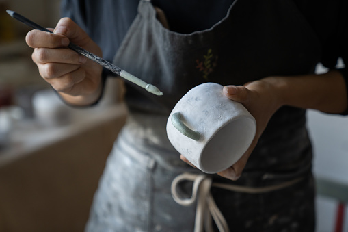 Experienced woman in black apron enjoys working with handmade craft in pottery studio. Female artisan uses brush to paint white ceramic cup with creative pattern standing on blurred background closeup