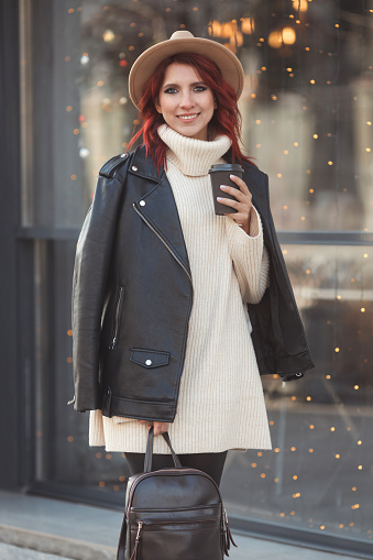 Vertical street style portrait of fashionable and stylish woman with red hair 25-30 years old in fashion casual clothes and holding take away hot coffee cup.