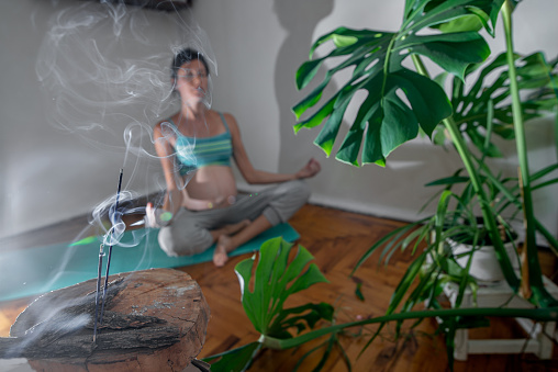 Pregnant woman sits on a yoga mat in a meditation pose. Pregnant woman doing yoga exercises at home. Incense room for relaxation during yoga. Yoga during pregnancy.