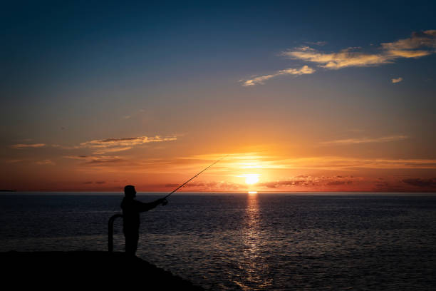 A silhouette of a man fishing off a rocky pier.
