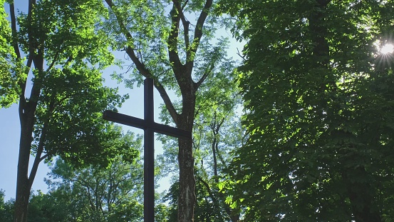 Tall Wooden Christian Catholic Crucifix Standing Among Trees in Church Park with Sun Rays Passing Through Leaves