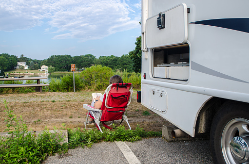 Woman reading in chair besides motorhome in public park with lake during summer day