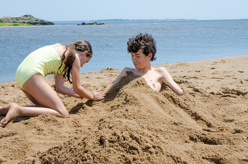 Teenage girl burying brother in beach sand during summer day vacations