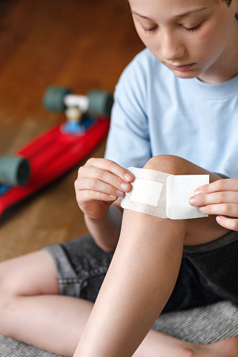 Boy putting sticking plaster on injured knee skin by himself. First aid for wounds.