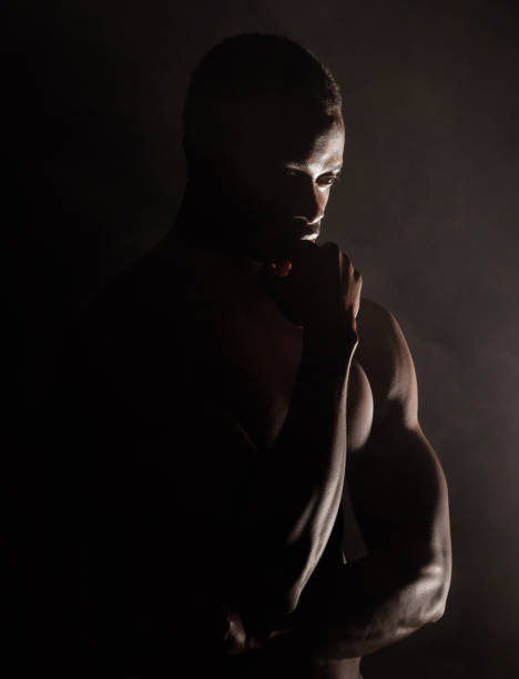 ein gutaussehender, muskulöser junger afroamerikaner im studio vor dunklem hintergrund. ein machohafter männlicher athlet, der nachdenklich aussieht, isoliert auf schwarz. bewegung von körper und geist. eine frage der psychischen gesundheit - chiaroscuro stock-fotos und bilder