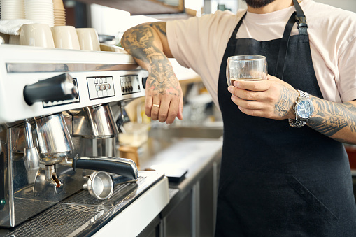 Cropped photo of barista in black apron leaning on coffee machine while holding a cup of espesso