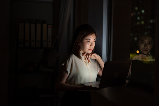 Young Asian business woman busy with office work on laptop at her work desk. She working late hour to complete her project reports before the deadline.