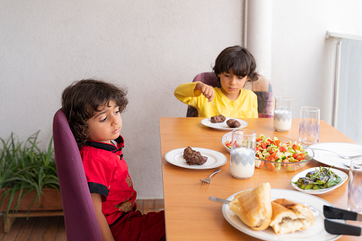 A table is set on the balcony of the house and meatballs are cooked on the barbecue. two boys with long hair and messy hair are sitting at the table waiting for food. The happy family photo was taken with a full frame camera in daylight.