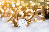 The golden watch lies on the snow against the background of the golden numbers 2023 with a golden defocus light