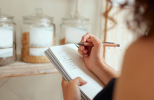 Budget planning, making shopping list and managing household expenses to save money. Financial accountability at home. Woman making shopping list for groceries on a notebook to plan a meal for dinner