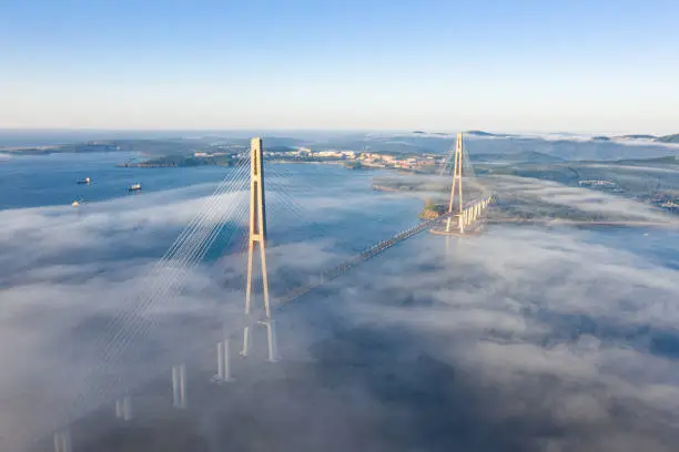 The pylons of the giant cable-stayed Russian Bridge rise above the seaside morning fog over the sea. Eastern Bosphorus Strait in the East Sea. View from a drone in 4k