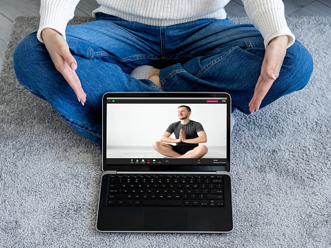 Online yoga class. Video chat. Virtual exercise. Web meditation program. Woman showing peaceful man sitting cross-legged with namaste position on laptop screen.