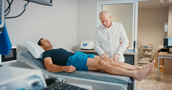 Physiotherapist doctor with patient in physical therapy for legs or muscle pain in a hospital exam consultation. Senior healthcare professional doing examination of of sports man on orthopedic table