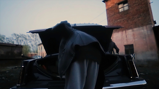Rear view of man in the leather jacket pulls the gun out of the trunk of the car. Man pulls out a riffle from the trunk of the car.