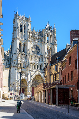 Amiens, France - August 13, 2022: The gothic facade of Notre-Dame d'Amiens cathedral with an old half-timbered townhouse in the foreground seen from a paved street on a sunny summer day.