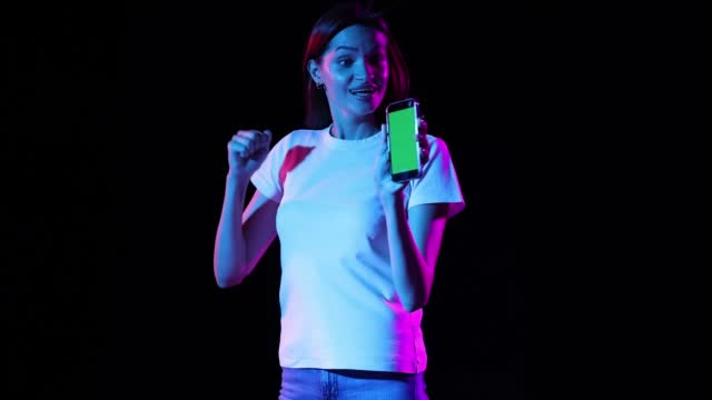 Young excited girl scrolling phone screen and shouting with delight isolated over dark background in neon light.