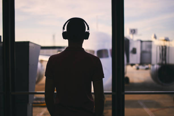 Silhouette of man with headphones while waiting for flight. Traveler looking from window of airport terminal before boarding to airplane.