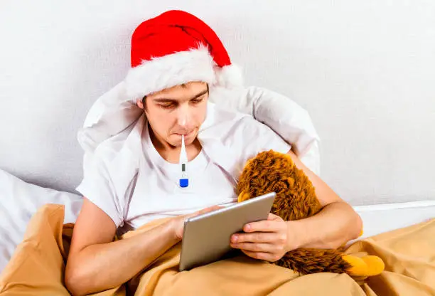 Sick Young Man in Santa Hat with a Thermometer and a Tablet Computer in the Bed