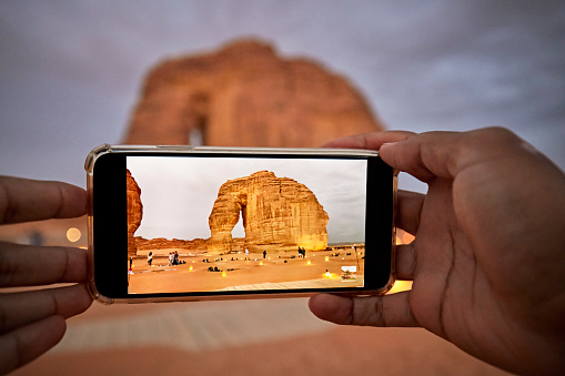 Focus on foreground device screen capturing a memory of red sandstone monolith shaped by wind and water erosion over millions of years. Al-Ula, Saudi Arabia.
