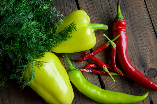 Different types of pepper and dill on the table. On a wooden table are two bell peppers, hot green peppers, hot red peppers and many small chillies with dill.