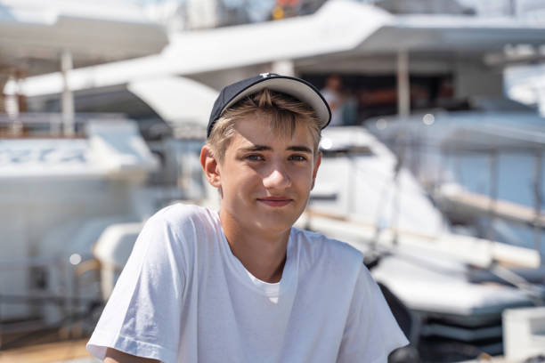 handsome teenager guy 16-18 years old smiling and looking at the camera over of the marina background. teenage boy dreams of traveling by sea on sailing ships. close up emotional portrait of  young man - years 13 14 years teenager old imagens e fotografias de stock