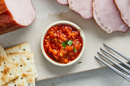 Hot salsa in a small handmade natural ceramic bowl, with ham and bread on a marble cutting board, served on a modern table, top view