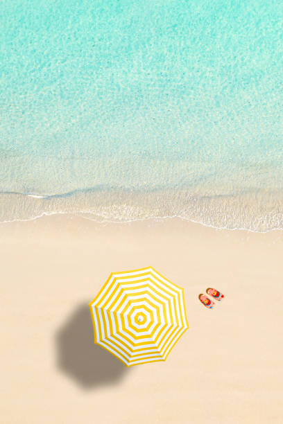 Aerial view of yellow umbrella, flip flops on sandy beach. Summer and travel concept. Blue, turquoise transparent water surface of ocean, sea, lagoon. Aerial, drone view. vertical stock photo