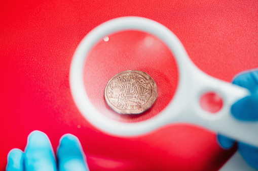 Hands holding a magnifying glass, examining an ancient coin.  Estimating value of a coin collection