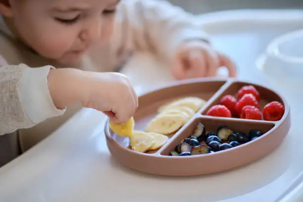 Photo of Toddler baby eats fruits and berries with his hand, table close-up. Child hands take food from a beige plate. Kid aged one year and two months