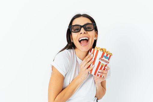 Cheerful young woman wearing 3D glasses, holding bucket of popcorn laughing, white studio background