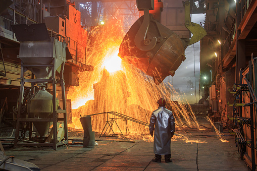 Hot steel pouring from big casting ladle into a mold in an iron foundry. Sparks, flame and smoke on the background. Factory worker controls the process.