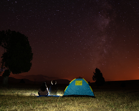 Photo of couple sitting next to camping tent and watching stars in the night. Shot with a full frame mirrorless camera.