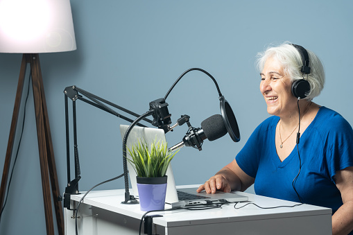 Photo of senior cheerful woman with white hair radio broadcasting via internet. She is wearing a blue blouse and using a laptop computer for podcasting. Shot with a full frame mirrorless camera.