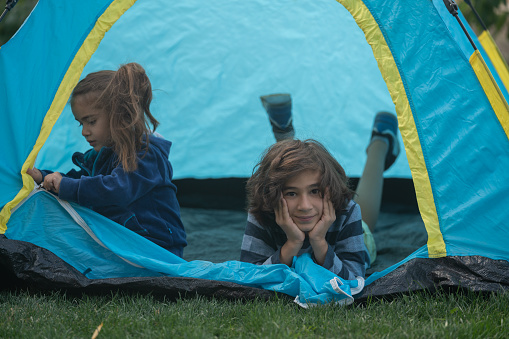Photo of 4 years old girl and 9 yers old boy posing in camping tent. They are brother and sister. The tent is turquoise colored. Shot under daylight with a full frame mirrorless camera.