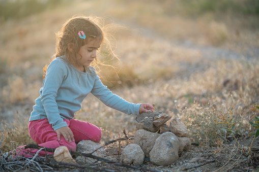 Photo of preschooler girl sitting on dirt and playing with small rocks and stones in nature. She is wearing a blue sweater and pink pants. Shot during sunset in outdoor with a full frame mirrorless camera.