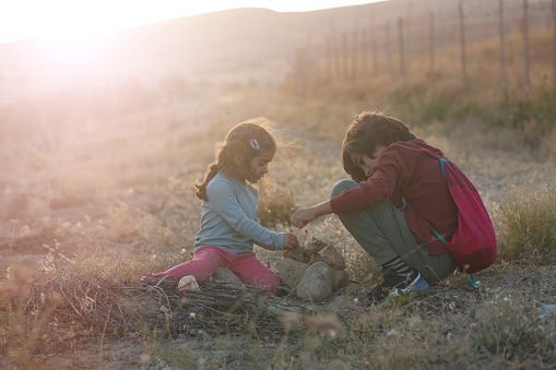 Photo of 4 years old girl and 9 years old boy playing with small stones and rocks in agricultural field. They are sitting on dirt. Shot under daylight during sunset with a full frame mirrorless. camera.