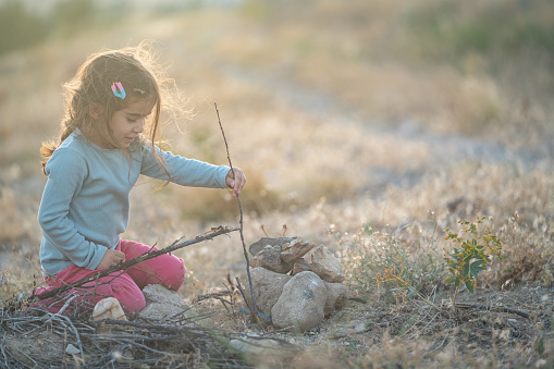 Photo of preschooler girl sitting on dirt and playing with small rocks and stones in nature. She is wearing a blue sweater and pink pants. Shot during sunset in outdoor with a full frame mirrorless camera.