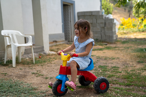 Photo of 4 years old girl riding multicolored tricycle in the backyard. She is wearing a blue dress. Shot under daylight with a full frame mirrorless camera.