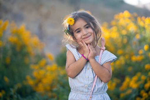 Portrait of 5 years old girl posing in front of yellow daisy flowers. She is wearing a striped blue dress. Shot under daylight with a full frame mirrorless camera.
