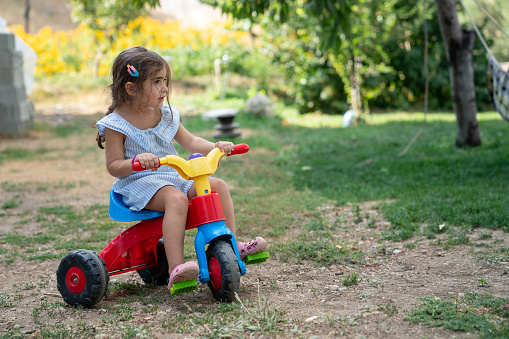 Photo of 4 years old girl riding multicolored tricycle in the backyard. She is wearing a blue dress. Shot under daylight with a full frame mirrorless camera.