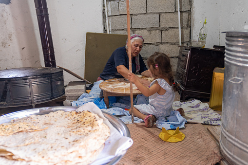 Candid photo of senior grandmother and 4 years old granddaughter baking traditional thin bread called yufka. They are in village house. Shot indoor with. a full frame mirrorless camera.