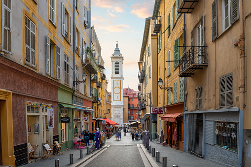 One of the picturesque narrow alleys and streets of shops and cafes in the Old Town Vieux Nice district of the city of Nice, France, on the Cote d'Azur French Riviera along the Mediterranean Sea. The bell tower of the Nice Cathedral in view at Place Rosetti.