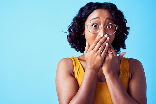 Money savings, discount sale, and deal surprised young teenage woman covering mouth with wow shock on blue background. Girl customer shopping with glasses scared by retail store news announcement.