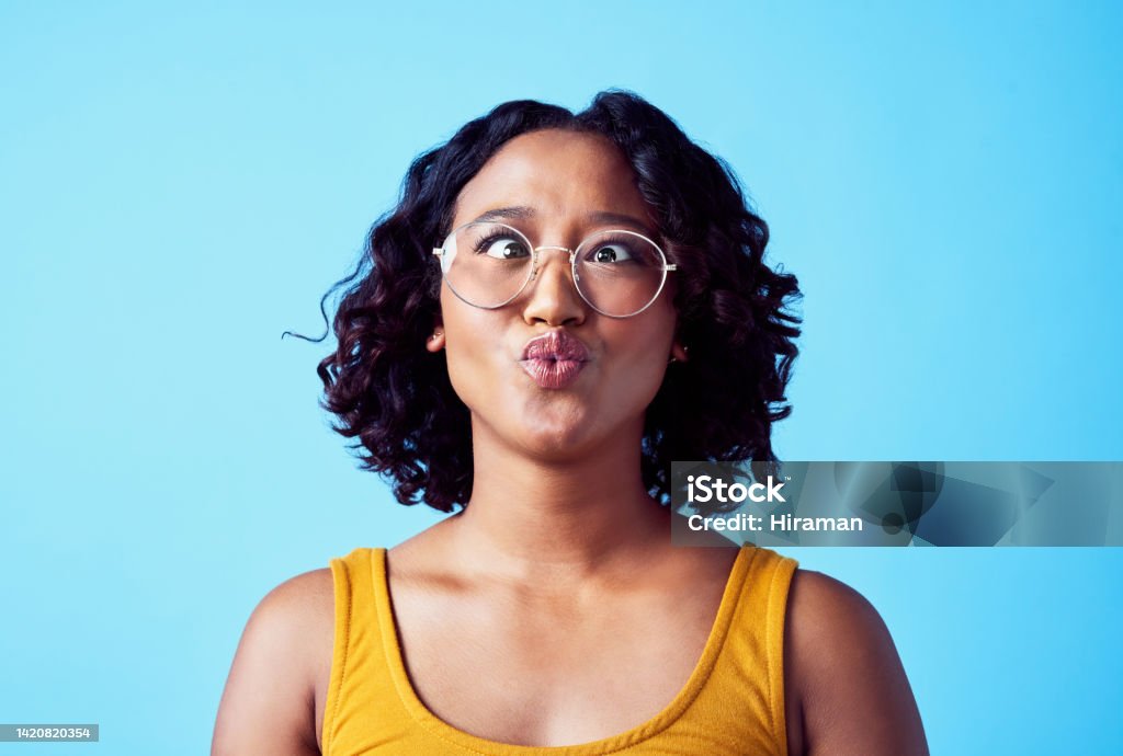 Crazy, comic and funny woman wearing glasses and making a silly face with cross eyed emoji against a blue background. Weird, fun and fool face expressions of a goofy girl with humour being awkward African Ethnicity Stock Photo