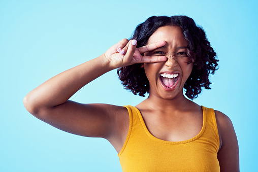 Happy black woman with a smile and a peace sign over her eye, with a blue background. Girl with her hand up to her face in celebration women, beauty and fun. Excited about love, life and her dreams.