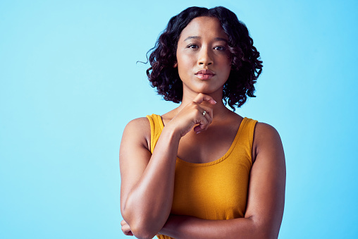 Portrait of a young black woman touching her face while standing in a studio with copy space. Beautiful, serious and mysterious African model thinking of an idea alone with a blue background.