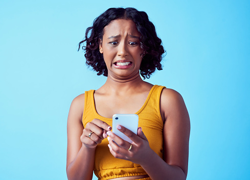 Scared mistake and error woman with smartphone, emoji expression and communication reading wrong email with studio blue background. Girl looking for unsend button on social media internet or tech app