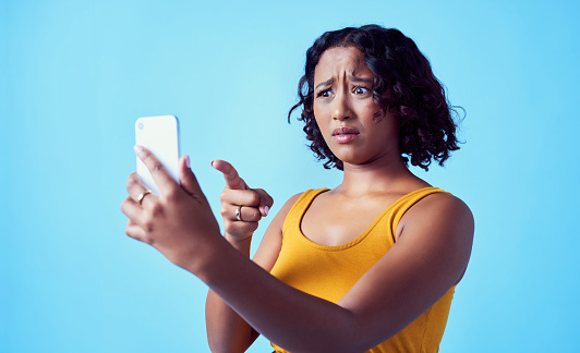 Social media, confused and phone of a woman pointing at smartphone and puzzled about screen display. Female having problems or trouble with mobile technology against a blue studio background.
