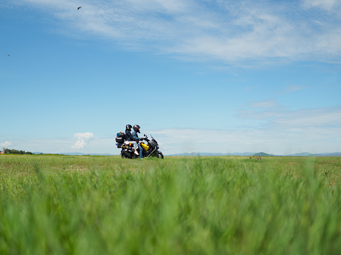 Young Asian couple sits on touring motorbike carrying equipment for long weekend camping trip. In the green meadow